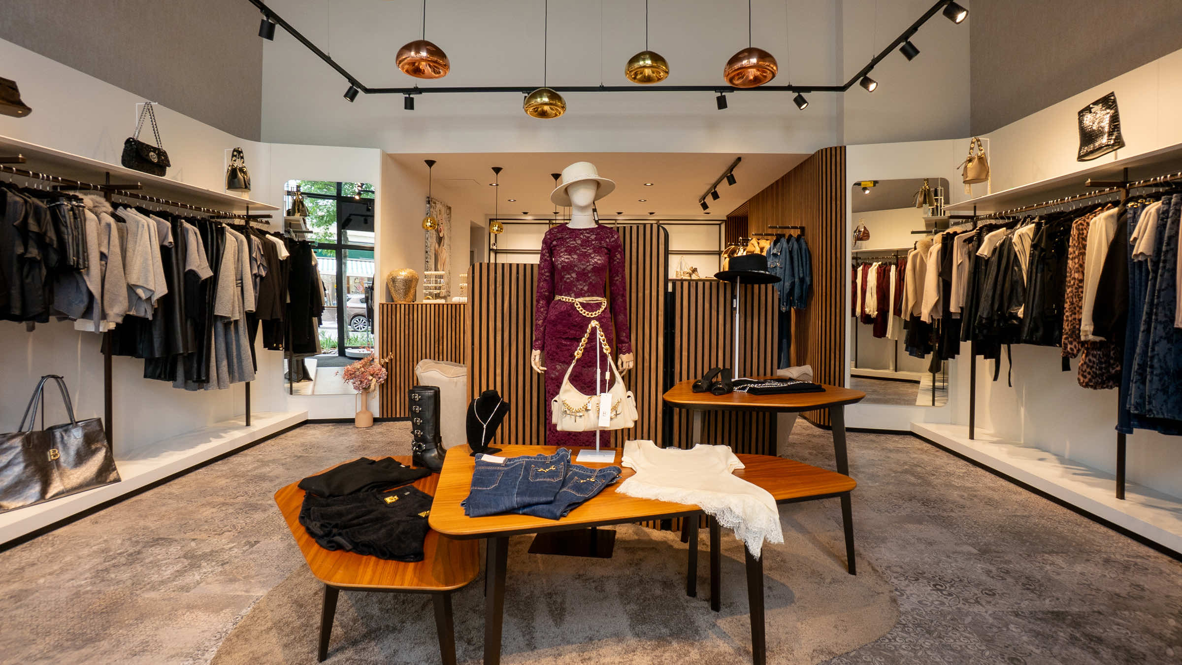 Verkaufsraum der LIMAA Boutique in Baden mit einer Schaufensterpuppe im bordeauxroten Spitzenkleid und Holztischen mit Jeans, Strick und Taschen