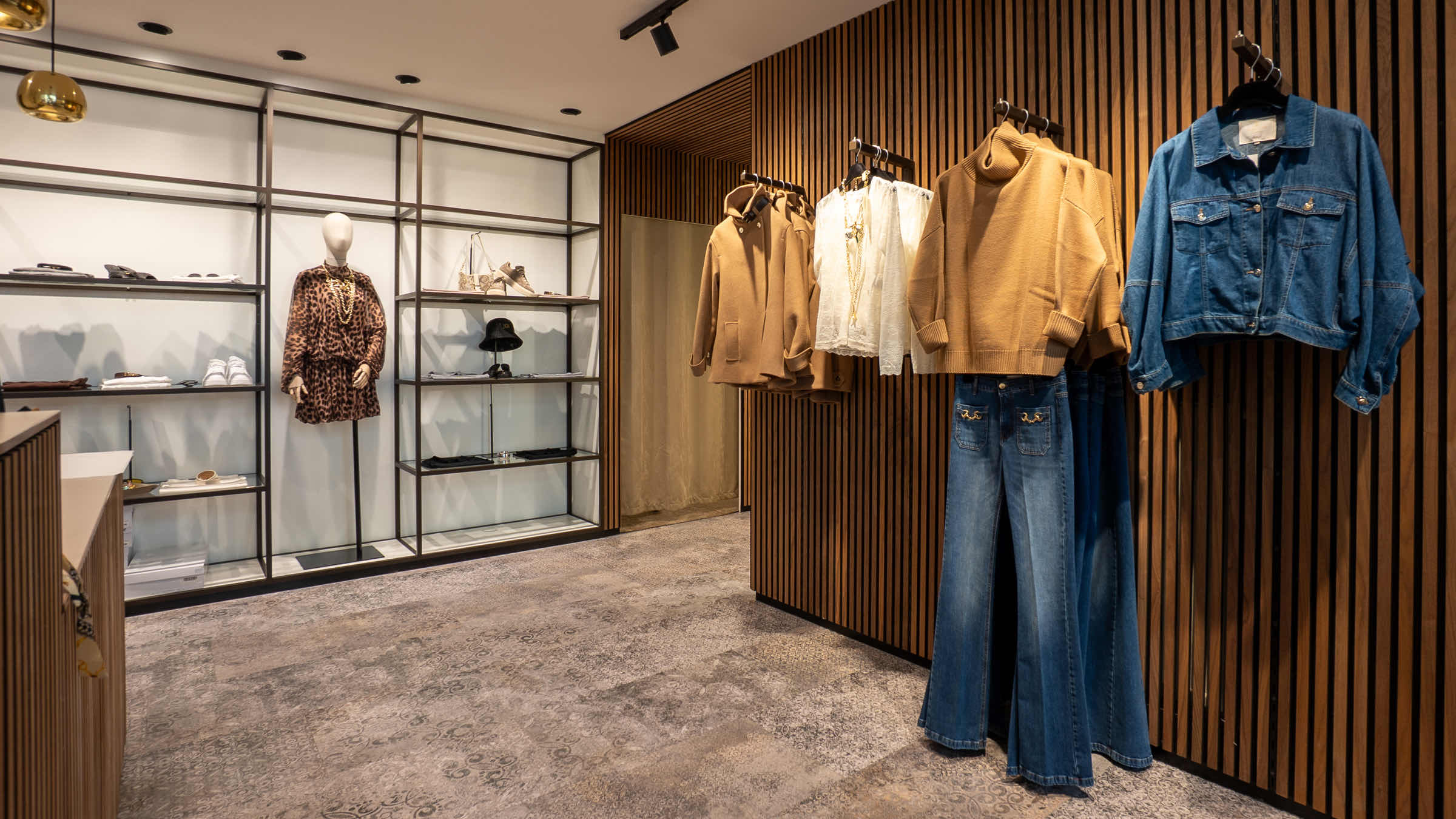 Outfit-Präsentation in Baden an einer Holzlamellenwand — Kamelmantel, Strickpullover, Jeansjacke und Schlaghose, daneben ein Regal mit Schuhen