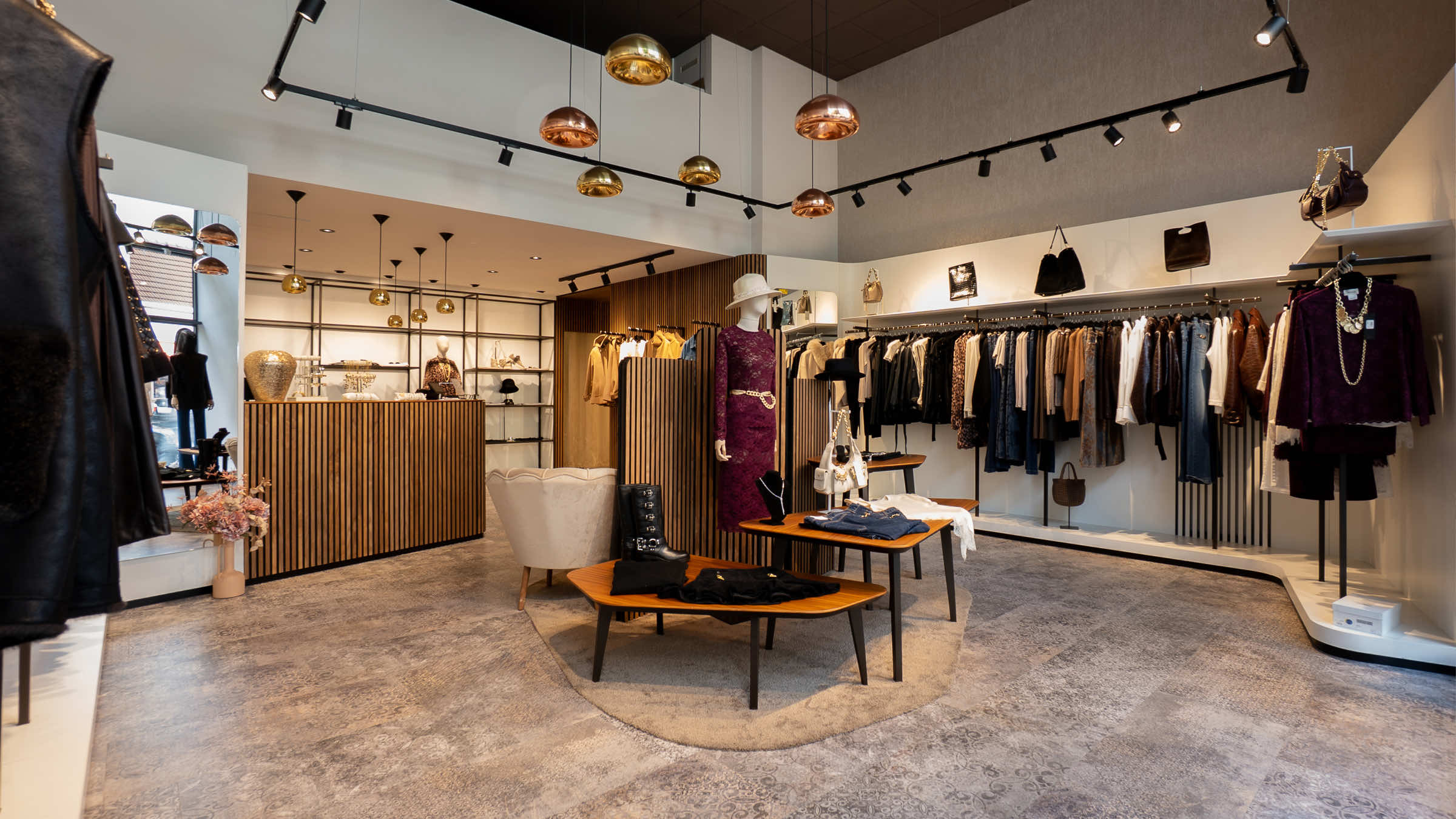 Weiter Überblick über die LIMAA Boutique in Baden mit Kupfer- und Goldleuchten, Holztresen, Kleiderstangen und mittig platzierten Sitzmöbeln