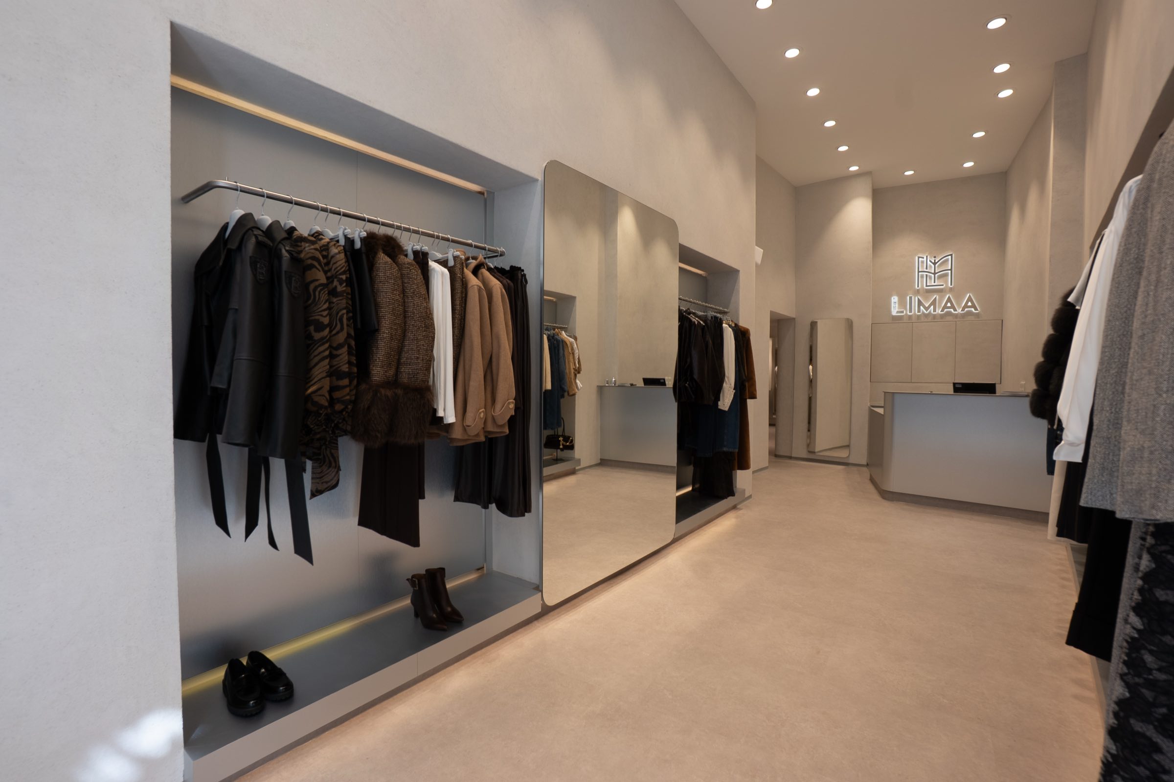 Innenraum der LIMAA Boutique in Wien (Tuchlauben): minimalistische Verkaufsfläche in warmen Beigetönen mit beleuchteter Kleider-Nische, großem Spiegel und dem LIMAA-Leuchtlogo über dem Tresen