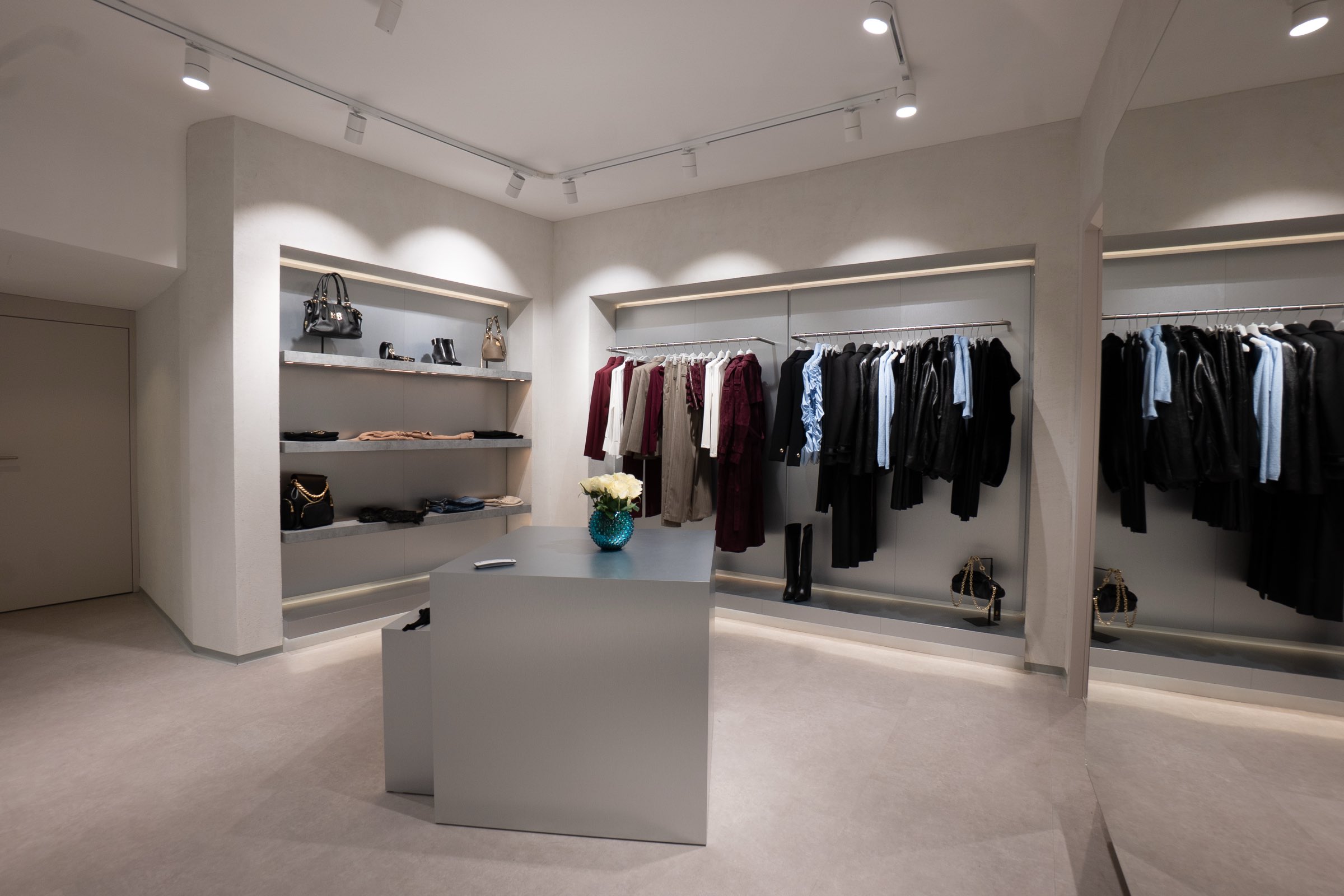 Verkaufsraum der Wiener LIMAA Boutique mit beleuchteten Wandregalen für Taschen und Stiefel, Kleiderstangen und einem grauen Mitteltisch mit weißen Rosen