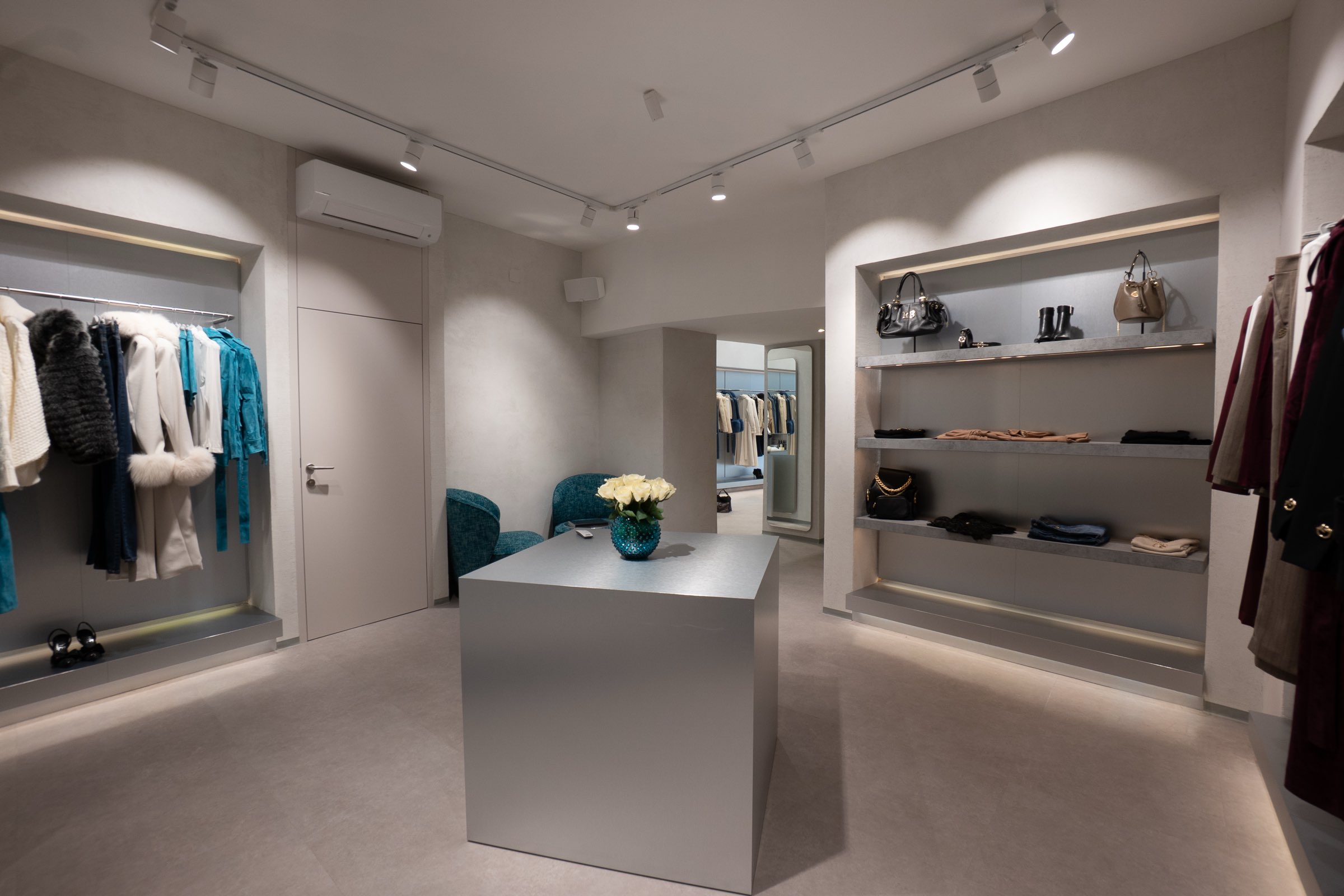 Überblick über die Wiener LIMAA Boutique mit Kleiderstange, beleuchteten Regalnischen für Taschen und Schuhe, grauem Mitteltisch mit Rosen und petrolfarbenen Sesseln